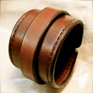 May include: A brown leather cuff bracelet with a double layer design. The bracelet is stitched with a contrasting thread.