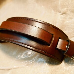 May include: A brown leather watch band with a silver buckle. The band has a unique design with a strap that goes over the top of the wrist.