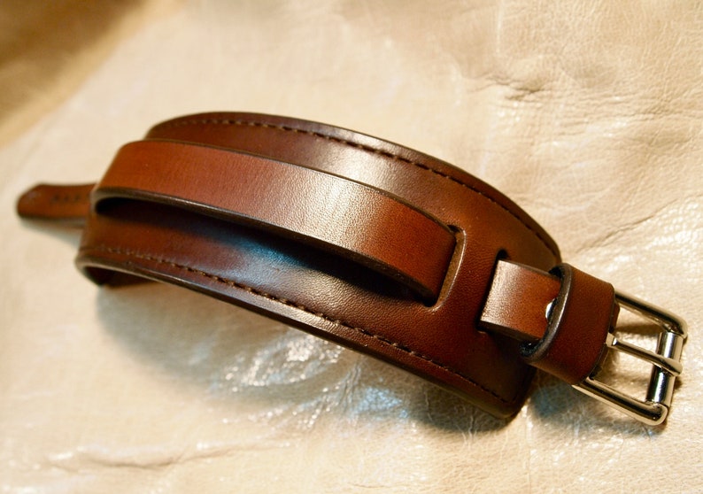 May include: A brown leather watch band with a silver buckle. The band has a unique design with a strap that goes over the top of the wrist.
