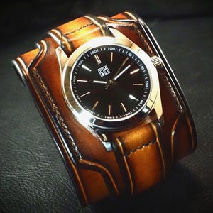 May include: A wristwatch featuring a round black analog face with silver-tone hands and markers, set within a wide, tan leather cuff strap. The leather band displays dark brown burnished edges and visible stitching, creating a layered appearance.