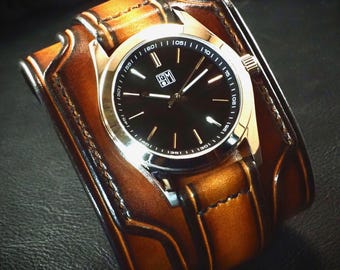 May include: A wristwatch featuring a round black analog face with silver-tone hands and markers, set within a wide, tan leather cuff strap. The leather band displays dark brown burnished edges and visible stitching, creating a layered appearance.