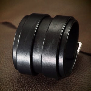May include: A wide, black leather cuff bracelet. The bracelet is made of several stacked bands of leather, with a silver-colored metal buckle closure. The cuff is shown on a brown leather surface.