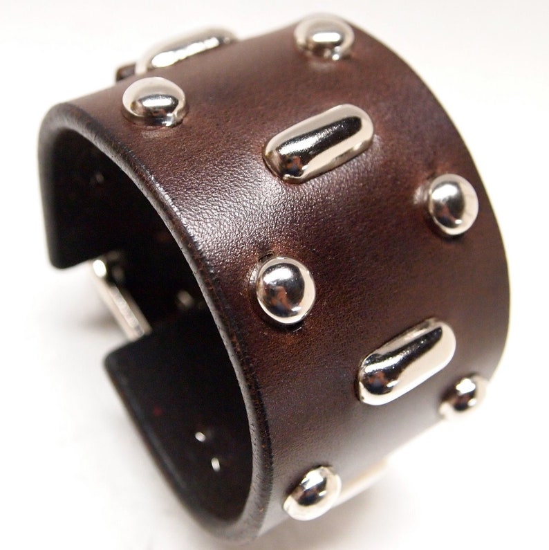 May include: A brown leather cuff bracelet with silver studs and metal accents. The bracelet has a buckle closure.