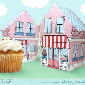May include: A printable kit for a sweet shoppe, featuring two pink houses with blue and white striped awnings. The houses have windows and doors, and the shoppe has a red and white striped awning. There is a cupcake with white frosting and sprinkles in the foreground.