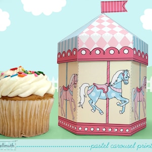 May include: A pastel pink and blue printable carousel party favour box with a white horse and a pink horse. The box is decorated with a pink and white striped flag on top. A cupcake with white frosting and colourful sprinkles is in the foreground.