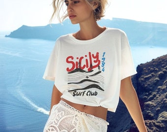 May include: A woman wearing a white t-shirt with the text "Sicily 1984 Surf Club" in red and blue. She is also wearing a white crochet skirt.