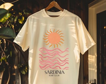 May include: A cream-colored t-shirt with a graphic of a sun setting over wavy pink and orange water. The text "Sardinia Surf Club" is printed below the graphic.