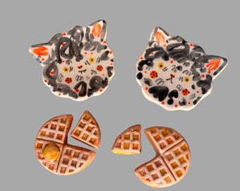 May include: Two ceramic cat-shaped pins with black and orange painted details, alongside two ceramic waffle-shaped pins. The cat pins feature abstract black markings and orange ears. The waffle pins are tan with grid patterns and orange accents.