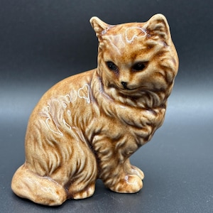 May include: A ceramic cat figurine in shades of brown. The cat is sitting with its front paws together and its head turned slightly to the side. The figurine has a glossy finish and detailed fur texture, with dark eyes and a small nose.