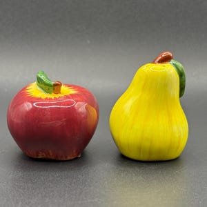 May include: A pair of ceramic fruit figurines. One is a red apple with a green stem and leaf, the other is a yellow pear with a green leaf and brown stem. The figurines are on a dark gray surface.