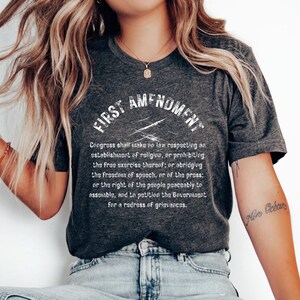 May include: A charcoal grey t-shirt featuring the words "FIRST AMENDMENT" in a white, distressed font. The full text of the First Amendment is printed below. The shirt is worn with light-wash denim jeans.