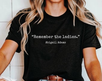 Pode incluir: Uma camiseta preta com texto branco que diz "Remember the ladies." O texto está em uma fonte de máquina de escrever e é atribuído a Abigail Adams.