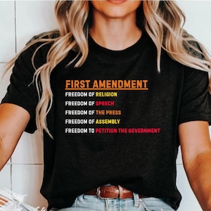 May include: A black t-shirt with the text "FIRST AMENDMENT" and a list of the five freedoms guaranteed by the First Amendment: Freedom of Religion, Freedom of Speech, Freedom of the Press, Freedom of Assembly, and Freedom to Petition the Government.