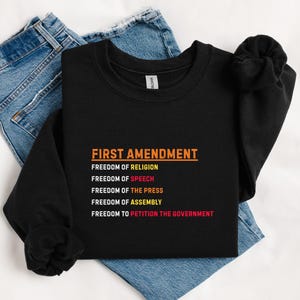 May include: Black sweatshirt with the text "FIRST AMENDMENT" in orange, and the freedoms of religion, speech, the press, assembly, and to petition the government listed below in white and red. The sweatshirt is laid on top of blue jeans.