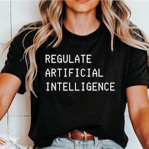 Puede incluir: Una camiseta negra de manga corta con el texto blanco "REGULATE ARTIFICIAL INTELLIGENCE" impreso en una fuente sans-serif clara en el pecho. La prenda tiene un cuello redondo clásico y un corte estándar con un diseño sencillo.