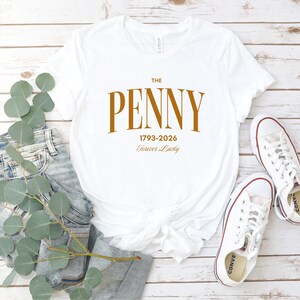 May include: White t-shirt with the text "THE PENNY 1793-2026 Forever Lucky" in a brown font. The shirt is paired with blue jeans and white sneakers with red accents. The items are arranged on a light wood-grain surface.