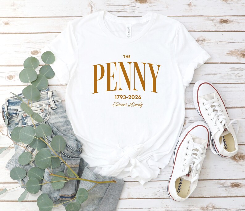 May include: White t-shirt with the text "THE PENNY 1793-2026 Forever Lucky" in a brown font. The shirt is paired with blue jeans and white sneakers with red accents. The items are arranged on a light wood-grain surface.