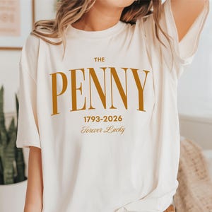 May include: Cream-colored t-shirt with the text "THE PENNY" in a bold, gold font. Below the text are the dates "1793-2026" and the phrase "Forever Lucky" in a script font. The shirt has a relaxed fit.