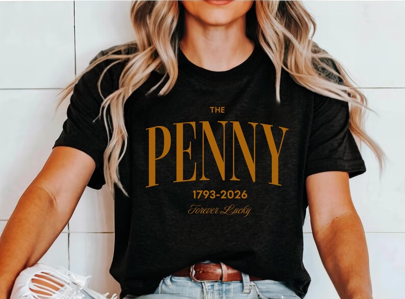May include: Black t-shirt with the text "THE PENNY" in gold lettering. Below the name are the dates "1793-2026" and the words "Forever Lucky". The shirt is worn by a person with long blonde hair.