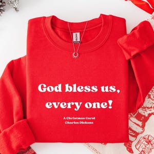 May include: A bright red sweatshirt with the quote "God bless us, every one!" in white text. The text "A Christmas Carol, Charles Dickens" is below the quote. A silver necklace with a sunburst pendant hangs from the collar.