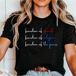 K&ouml;nnte beinhalten: Schwarzes T-Shirt mit dem Text "freedom of speech", "freedom of religion" und "freedom of the press" in wei&szlig; mit roten und blauen Akzenten.