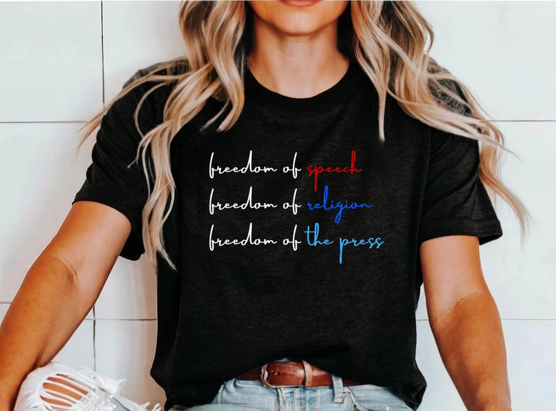 K&ouml;nnte beinhalten: Schwarzes T-Shirt mit dem Text "freedom of speech", "freedom of religion" und "freedom of the press" in wei&szlig; mit roten und blauen Akzenten.