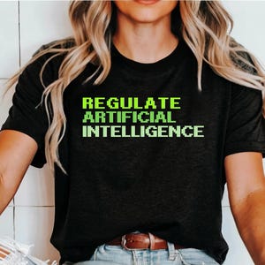 Może przedstawiać: Czarny t-shirt z krótkim rękawem z napisem "REGULATE ARTIFICIAL INTELLIGENCE" nadrukowanym jaskrawą, limonkową, pikselową czcionką na klatce piersiowej. Koszulka ma okrągły dekolt i standardowy krój wykonany z jednolitego czarnego materiału.