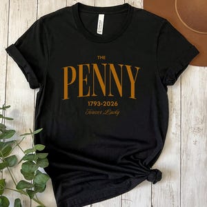 May include: Black t-shirt with the text "THE PENNY 1793-2026 Forever Lucky" in a gold-colored font. The shirt is made of a soft material and has a crew neck. The sleeves are rolled up.