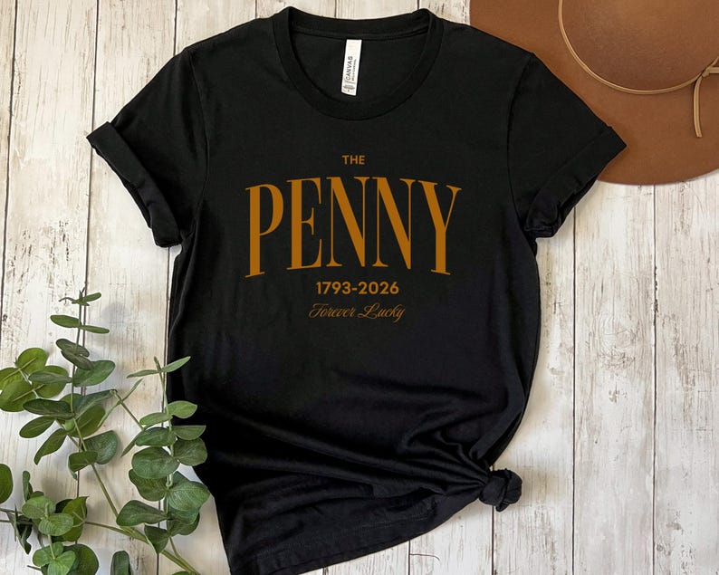 May include: Black t-shirt with the text "THE PENNY 1793-2026 Forever Lucky" in a gold-colored font. The shirt is made of a soft material and has a crew neck. The sleeves are rolled up.