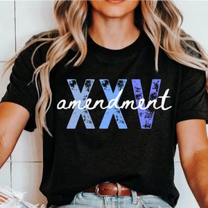 May include: Black t-shirt featuring the text "XXV amendment" in a blue and purple gradient. The word "amendment" is in a white script font. The shirt has a crew neck and short sleeves.