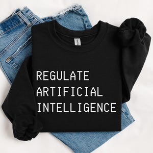 May include: A black long-sleeve sweatshirt featuring the white text "REGULATE ARTIFICIAL INTELLIGENCE" printed in a clean, sans-serif font on the chest, paired with a pair of blue denim jeans folded underneath the garment.