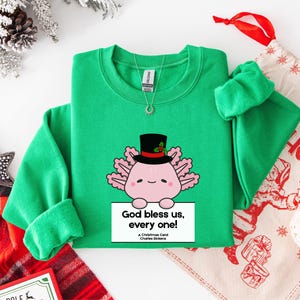 May include: A green sweatshirt featuring a cartoon axolotl wearing a top hat and holding a sign that reads "God bless us, every one!" from A Christmas Carol. The design is festive and holiday-themed, with a Christmas stocking and decorations in the background.