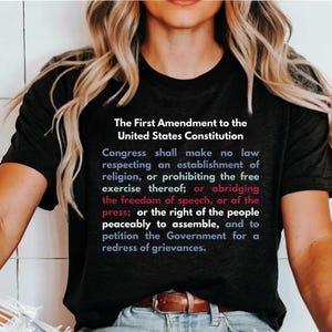 May include: A black t-shirt with the text "The First Amendment to the United States Constitution" and the full text of the First Amendment printed in red, white, and blue.