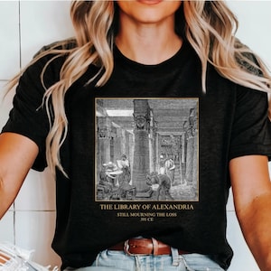 May include: Black t-shirt featuring a detailed black and white illustration of the Library of Alexandria. The image includes the text "THE LIBRARY OF ALEXANDRIA STILL MOURNING THE LOSS 391 CE".