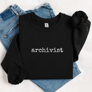 May include: Black sweatshirt with the word "archivist" printed in white, resting on a pair of blue jeans. The sweatshirt has long sleeves and a crew neck. The jeans are folded beneath the sweatshirt.