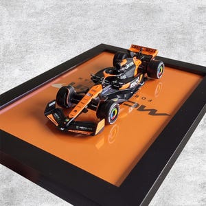May include: A framed model Formula 1 race car, predominantly black and orange, with sponsor logos. The car sits on an orange background within a black frame. The wheels have colorful accents.