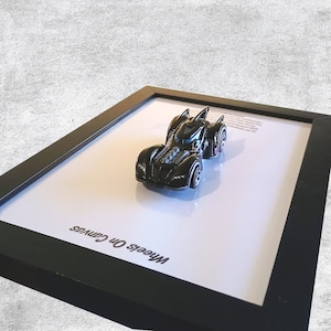 Puede incluir: Un coche de juguete negro con un diseño futurista, presentado en un marco negro sobre un fondo blanco. El coche tiene detalles azules y plateados y está colocado sobre una superficie blanca. El marco tiene el texto "Wheels On Canvas".