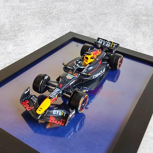Peut inclure: Maquette détaillée d'une voiture de course de Formule 1, principalement noire avec des accents rouges et jaunes. La voiture présente des logos de sponsors tels que "BYBIT" et "Oracle". Le modèle est encadré en noir, sur fond bleu.