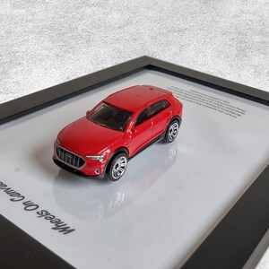 以下が含まれることがあります： 黒いフレームに白いマットが敷かれた、赤いAudi Q4 e-tronのミニカーモデル。フレームの左下には「Wheels On Canvas」の文字が印刷されています。