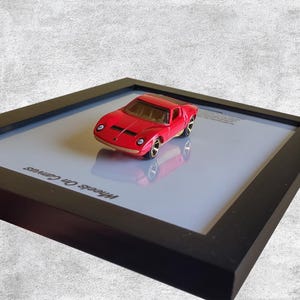 Könnte beinhalten: Ein rotes Spielzeugauto mit goldenen Rädern wird in einem schwarzen Rahmen präsentiert. Das Auto ist ein klassisches Sportwagendesign. Der Rahmen befindet sich auf einer hellblauen Oberfläche. Der Text "Wheels On Canvas" ist sichtbar.