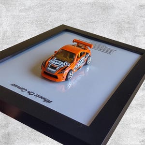 Może przedstawiać: Pomarańczowy plastikowy samochód z czarnymi i białymi paskami wyścigowymi i napisem "Alien" na boku. Samochód jest oprawiony w ramę i prezentowany na białym tle z napisem "Wheels on Canvas".