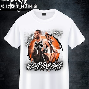 May include: White t-shirt featuring a graphic of a basketball player in a black and white uniform with the name "WEMBANYAMA" in a stylized font. The shirt also includes the text "VICTOR" and the number "1".