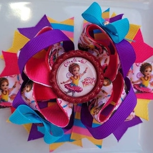 May include: A colorful hair bow with a red, white, and blue design. The center of the bow features a round, red button with a picture of a girl in a pink dress and the words "C'est la la!" and "Fancy Nancy".