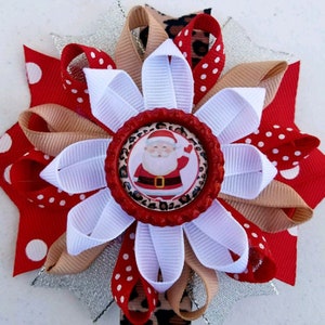 Peut inclure: Un nœud de cheveux de Noël festif avec une image du Père Noël au centre. Le nœud est composé de rubans rouges, blancs, beiges et léopard, avec un accent argenté scintillant. Le design est circulaire, en forme de fleur.