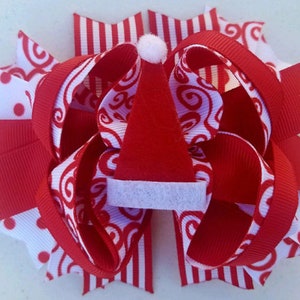 May include: Red and white Christmas hair bow with a red Santa hat in the center. The bow is made of three different ribbons: red with white polka dots, red with white stripes, and white with red swirls.