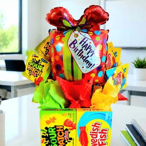 May include: A gift arrangement featuring a red balloon shaped like a present with a Happy Birthday tag, surrounded by Sour Patch Kids and Swedish Fish candy boxes and bags, accented with bright green, red, and yellow tissue paper filler.