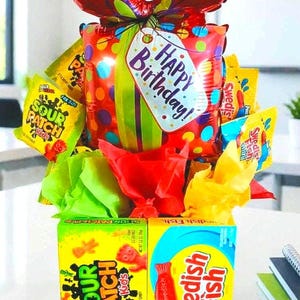 May include: A festive birthday gift arrangement featuring a red bow-shaped balloon, a balloon with colorful polka dots, and a tag that says "Happy Birthday!". The arrangement includes boxes of Sour Patch Kids and Swedish Fish candies, and colorful tissue paper.
