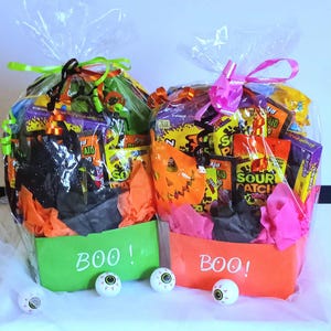 May include: Two Halloween gift baskets filled with candy and treats, wrapped in cellophane and tied with ribbons. One basket is green with "BOO!" written on it, the other is orange with "BOO!" written on it. Eyeball decorations are in front of the baskets.