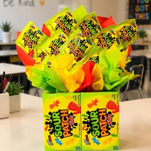 May include: A candy bouquet featuring multiple individual yellow Sour Patch Kids snack bags arranged with bright red, yellow, and green tissue paper. The base consists of two rectangular Sour Patch Kids boxes placed side by side on a flat surface.