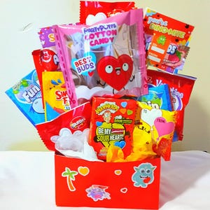May include: A red gift box filled with various candy packets, including cotton candy, sour candies, and Skittles. The box is decorated with stickers and the text "Be My Sour Heart!" is visible on one of the candy wrappers.
